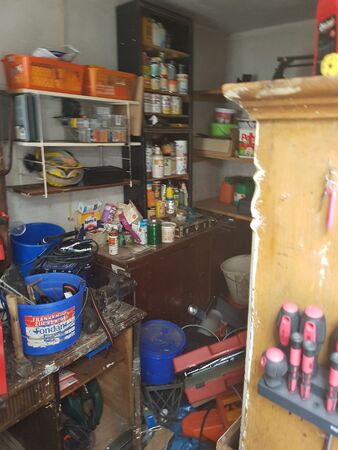 Unordentliche Werkstatt mit Werkzeug, Regalen, Farbeimern und verschiedenen Haushaltsgegenständen.